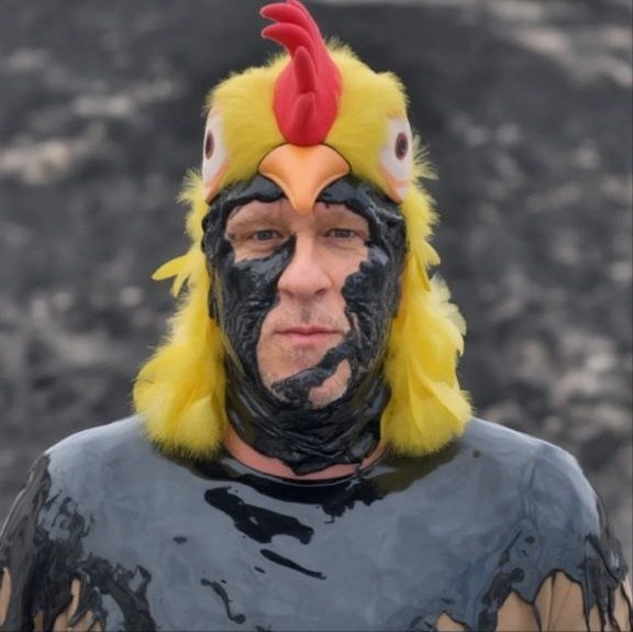 Portrait of a person wearing a yellow chicken costume headpiece and dark theatrical makeup.